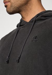 Άνδρας που φοράει μαύρο φούτερ Champion με κορδόνια και κεντημένο λογότυπο στο στήθος, με ορατό μέρος του προσώπου και του λαιμού του.