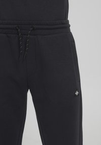Sorte joggebukser med elastisk midje, snor med detaljer, sidelommer og et lite logo-patch på venstre side.