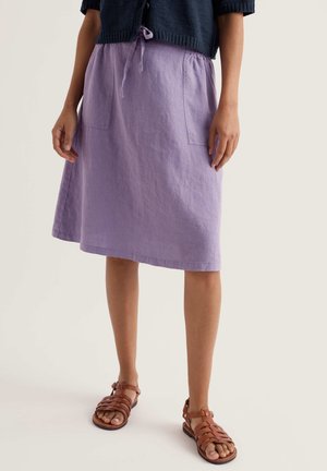 Frau trägt einen knielangen Lavendelfarbenen Rock mit Vordertaschen, einen marineblauen, kurz geschnittenen Pullover und braune Sandalen mit Riemchen, steht vor einem hellen Hintergrund.