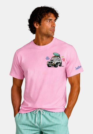 Uomo con capelli ricci indossa una maglietta rosa con grafica di un'auto dei cartoni animati e una tavola da surf e pantaloncini a strisce turchesi, in piedi con le mani nelle tasche.