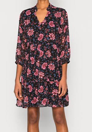 Frau trägt ein schwarzes Kleid mit pinkem und blauem Blumenmuster, dreiviertel Ärmeln und einem stufigen Rock.