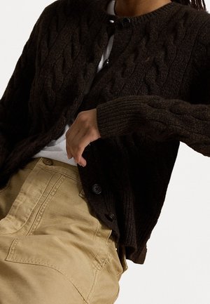 Persona che indossa un cardigan lavorato a trecce di colore marrone scuro sopra una maglietta bianca, abbinato a pantaloni beige a vita alta con cuciture visibili e dettagli con bottoni.