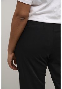 Černé kalhoty s upraveným střihem, které mají elegantní texturu a jednu zadní kapsu. Ruka modelky spočívá na bocích, což ukazuje na detail lokte.