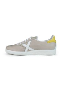 Zapatilla de piel beige con cordones blancos, suela de goma blanca, acentos verdes y amarillos, y detalles perforados en los laterales.