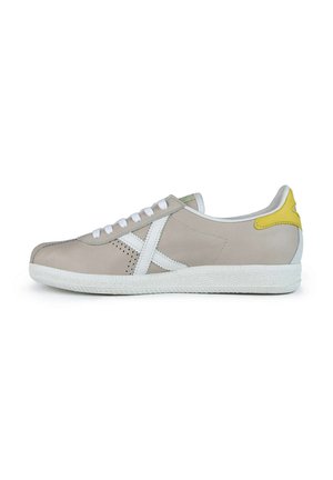 Zapatilla de piel beige con cordones blancos, suela de goma blanca, acentos verdes y amarillos, y detalles perforados en los laterales.