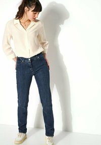 Blouse boutonnière crème clair à manches trois-quarts, associée à un jean slim bleu foncé. Mises en valeur par des baskets blanches et un collier délicat.