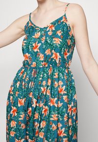 Kvinna klädd i en tealfärgad klänning med orange och vit blommig print samt tunna axelremmar, med ett knytbart midjebälte med fransar.