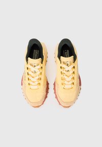 Pair of yellow Lacoste sneakers with beige suede accents and brown rubber soles, viewed from above on a white background.