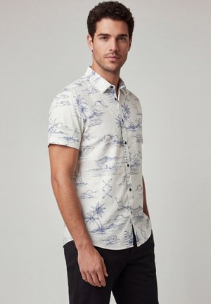 Man draagt een wit overhemd met korte mouwen met blauwe tropische print en zwarte broek, staand tegen een effen lichtgrijze achtergrond.