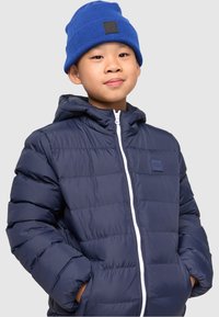 Marineblå puffjakke med hette, med en hvit glidelås og logo-lapp. Båret med en blå strikket lue. Glatt tekstur med quiltet design.