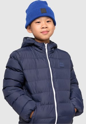 Giacca puffy blu navy con cappuccio, dotata di una cerniera bianca e un patch con logo. Indossata insieme a un berretto di maglia blu. Tessuto liscio con design trapuntato.