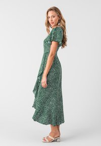 Vestido floral verde con mangas cortas, dobladillo con volantes, patrón de puntos blancos y un diseño fluido; combinado con sandalias de tacón bloque de color claro.