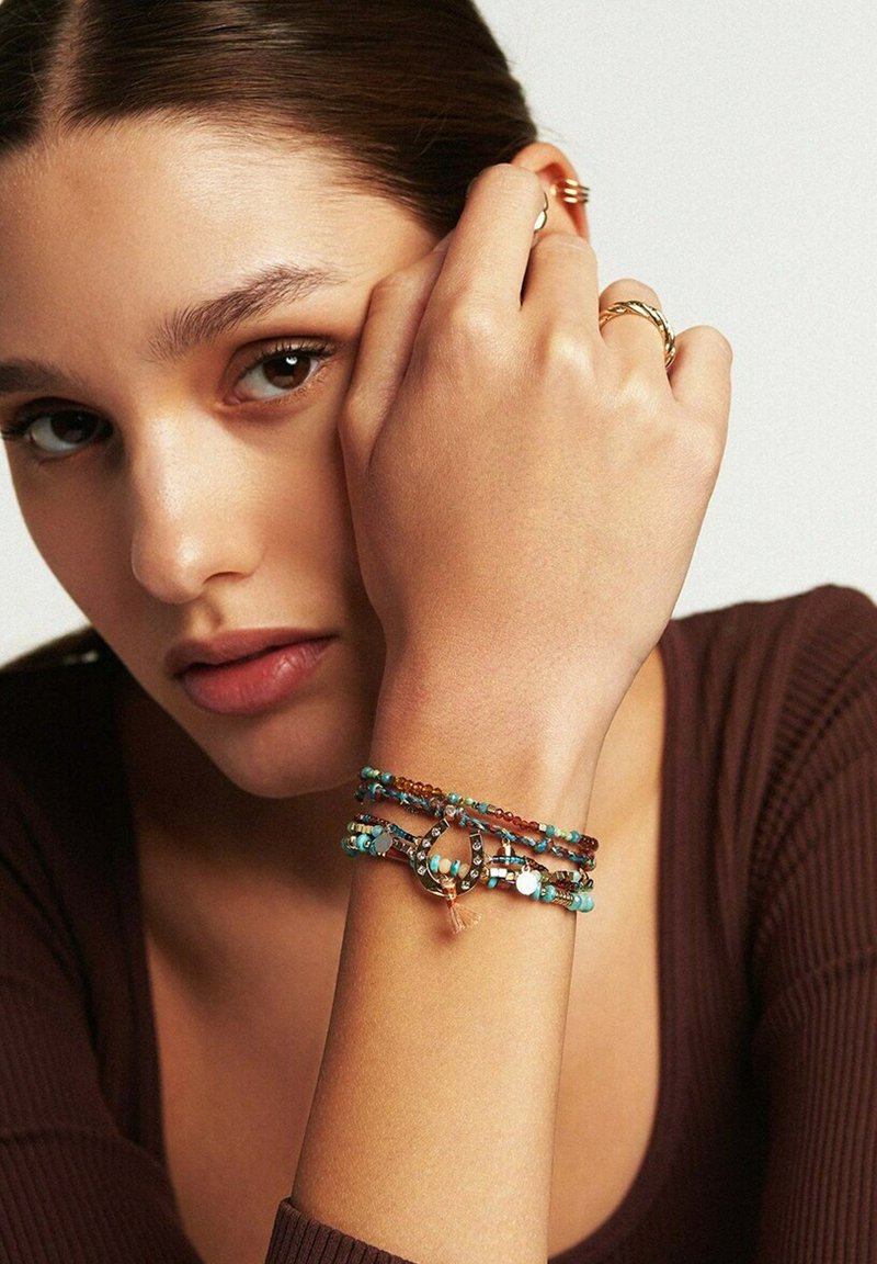 Bracelet en perles multicolores comprenant des accents turquoise, marron et argent, avec un détail de nœud décoratif. Porté au poignet.