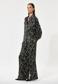 Črno vzorčasto dvodelno oblačilo, ki vključuje ohlapen srajčast gumb z dolgimi rokavi in široke hlače, z geometrijskim bež vzorcem in gladko teksturo.