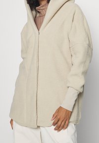 Beige fleecejacka med huva, med dragkedjestängning, oversize-passform och texturerad, mjuk yta. Inkluderar ribbade mudd och fåll.