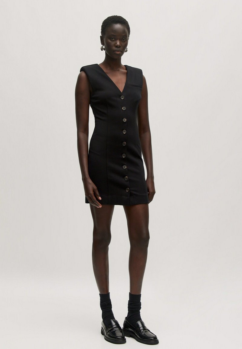 Schwarzes ärmelloses Minikleid mit V-Ausschnitt, vorderen Knöpfen und körpernaher Silhouette. Begleitet von knöchellangen Socken und Loafers.