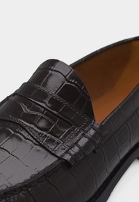 Γυαλιστερό μαύρο δερμάτινο μοκασίνι με υφή κροκοδείλου και λεπτομέρεια λουριού penny, που δείχνει κοντινή εικόνα του πάνω μέρους και της εσωτερικής επένδυσης.