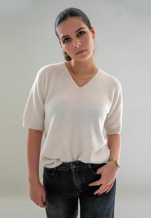 Witte, lichtgewicht gebreide trui met korte mouwen en een V-hals, gecombineerd met donkere, aangesloten jeans. Accessoires omvatten een gouden horloge en ringen.