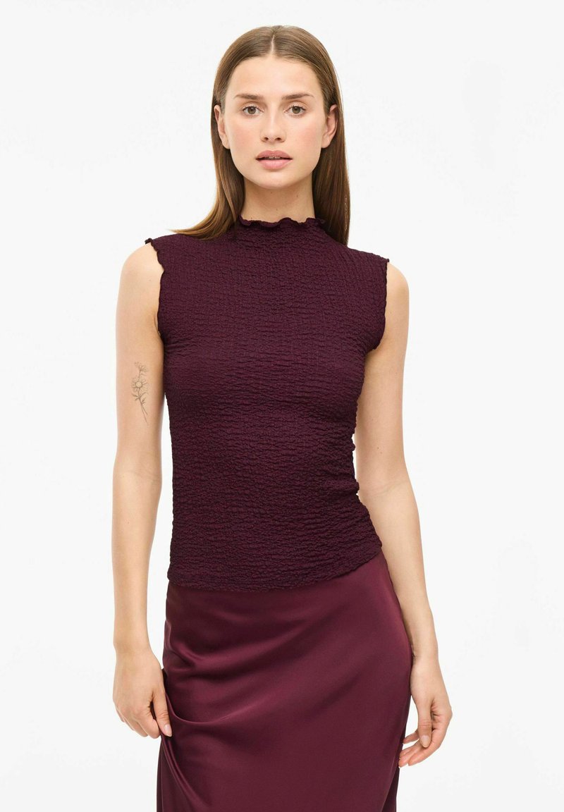 Burgunderfärgad ärmlös topp med en texturerad tyg, med en volangkantad hög krage och en figurnära design. Slät satinkjol i matchande färg.