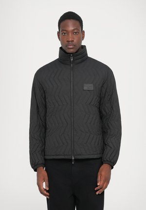 Mann in einer schwarzen gesteppte Jacke mit hohem Kragen und Reißverschluss, der vor einem einfachen weißen Hintergrund steht.