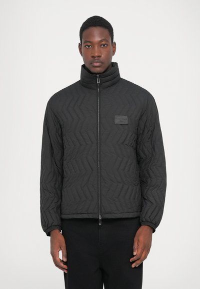 Homme portant une veste matelassée noire avec un col haut et une fermeture éclair à l'avant, se tenant devant un fond blanc uni.