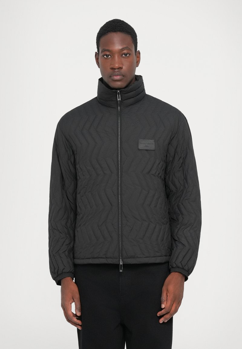 Man wearing a black quilted jacket with a high collar and zip front, standing against a plain white background.
