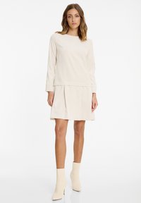 Eine Frau steht in einem cremefarbenen Langarmkleid mit plissierter Rockpartie und passenden cremefarbenen Stiefeletten auf einem weißen Hintergrund.