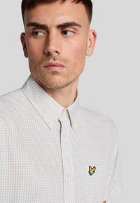 Camicia a maniche corte con colletto in un motivo a quadri beige chiaro e crema, con un piccolo logo giallo sul taschino sinistro. Tessuto strutturato.