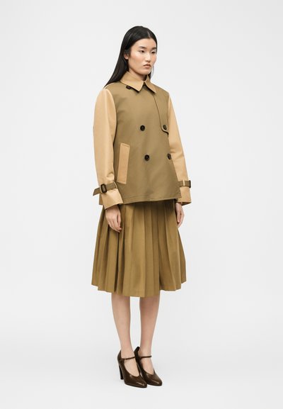 Γυναίκα που φοράει διχρωμο μπεζ και καφέ σακάκι διπλού πέτου με λεπτομέρειες με αγκράφες, πλισέ midi φούστα και καφέ ψηλοτάκουνα παπούτσια Mary Jane, στέκεται όρθια.