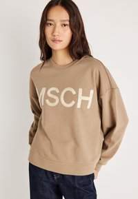 Καφέ φούτερ με λαιμόκοψη, χαμηλούς ώμους και έντονα, μαλακά λευκά γράμματα "MSCH" μπροστά. Φοριέται με σκούρα τζιν.