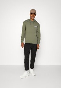 Homme portant un hoodie vert olive avec cordon de serrage, poche frontale et écusson logo. Pantalons noirs ajustés, baskets blanches et casquette beige. Fond uni.