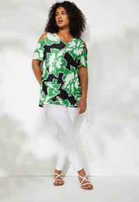 Top nero con motivo floreale verde, spalle scoperte e maniche corte, abbinato a pantaloni bianchi attillati e sandali bianchi con cinturino.
