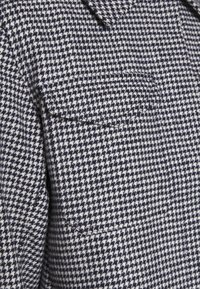 Kurtka z wełnianego materiału w wzór houndstooth w kolorze granatowym i białym. Posiada kieszeń na piersi oraz klasyczny design kołnierza. Teksturowane wykończenie.