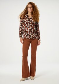 Leopardprint knoopblouse in tan, bruin en roze; gecombineerd met bruine flared corduroypants. Model draagt naakte haksandalen.