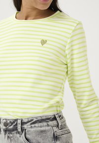 Lange mouw gestreepte top in lichtgroen en wit met een klein gouden hartembleem op de borst, gecombineerd met hoogdonkergrijze denimshorts.