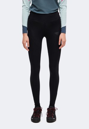 Persona che indossa leggings neri aderenti, scarpe da trekking nere con lacci rossi e una maglia blu-grigio a maniche lunghe, in piedi davanti a uno sfondo neutro.