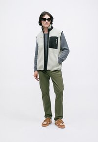 Persona con gafas de sol negras, suéter de mangas largas gris, chaleco de forro polar gris claro con bolsillo negro, pantalones oliva y zapatillas marrones, de pie.