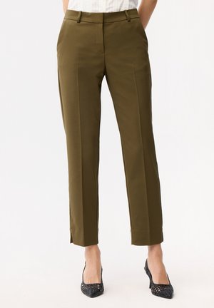 Personne portant un pantalon sur mesure olive vert longueur cheville et des chaussures à talons hauts noires tissées, debout les mains dans les poches.
