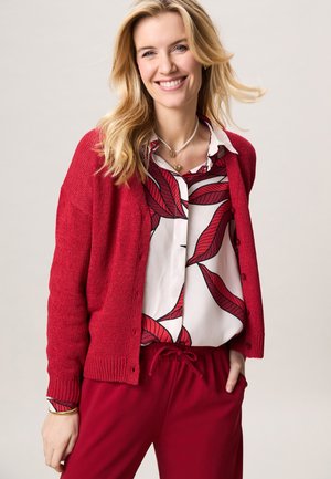 Vrouw die lacht, draagt een rode cardigan over een wit blouse met rode bladmotieven en een rode broek met koordje tegen een effen lichte achtergrond.