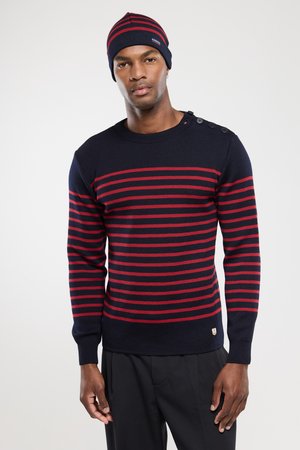 Man wearing navy blue and red striped sweater with buttons on shoulder and matching navy blue beanie against plain background.