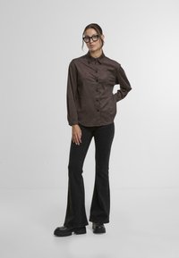 Brown, long-sleeve shirt with a button-down front and collar, paired with flared black pants and chunky black shoes. Smooth texture.