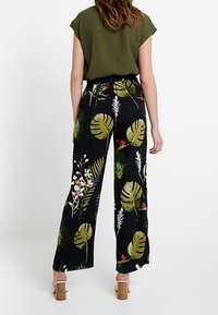 Svarta vida byxor med grön och vit tropisk blad- och blommönster. Matchas med en olivgrön topp med kortärm och blockklackar.