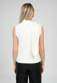 Femme aux longs cheveux blonds portant un pull sans manches blanc et un pantalon noir, vue de dos sur un fond uni.