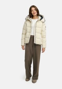 Veste en duvet de couleur beige clair, présentant une fermeture éclair sur le devant, une capuche surdimensionnée et un design matelassé. Tissu texturé avec une finition lisse.