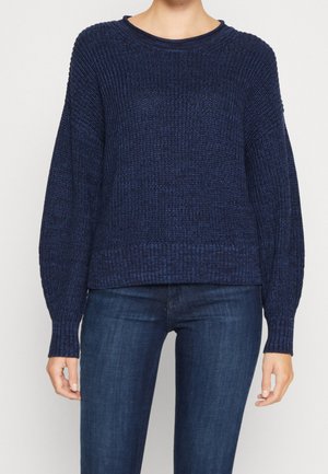 Kobieta ubrana w ciemnoniebieski, teksturowany sweter z długim rękawem i dopasowane, ciemnoniebieskie dżinsy, stojąca na jednolitym tle.