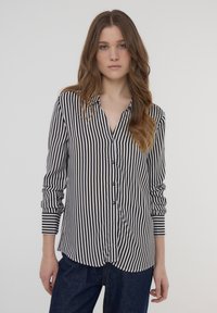 Camicia button-up a righe bianche e nere con maniche lunghe, dotata di colletto e orlo curvo. Tessuto morbido con superficie liscia.