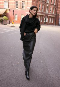 Schwarzer Strickpullover mit einem drapierten Ausschnitt, kombiniert mit einem hoch sitzenden schwarzen Lederrock und schwarzen Stiefeletten, auf einer Straße mit Backsteingebäuden.