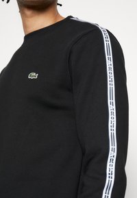 Černá mikina s kulatým výstřihem. Obsahuje malé logo krokodýla v zelené a bílé barvě a bílé pruhované nápis "LACOSTE" na rukávech.