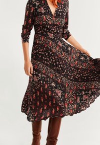 Robe noire avec des motifs paisley et floraux en rouge et crème. Manches longues, décolleté en V et jupe plissée. Associée à des bottes marron hauteur genou.