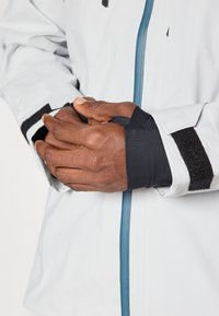 Chaqueta gris claro con un zipper azul, puños negros y detalle de Velcro, que presenta un primer plano de manos situadas cerca de la zona de la muñeca.